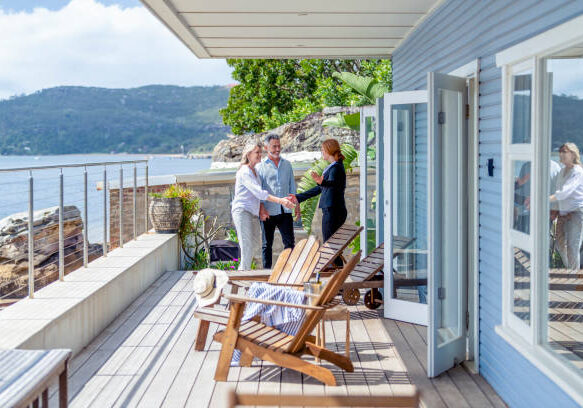 24-35 Real estate agent showing a mature couple a new house. The house is contemporary. All are happy and smiling and shaking hands. The couple are casually dressed and the agent is in a suit. Waterfront can be seen in the background